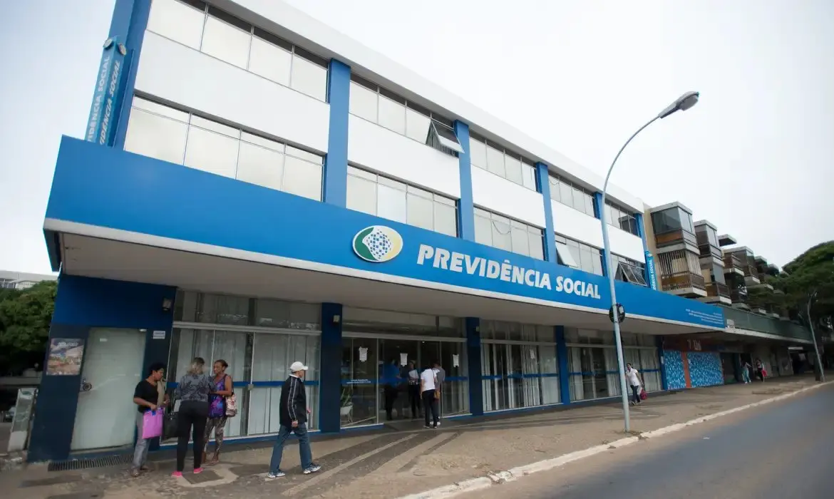 Fachada de uma agência da Previdência Social (INSS) com seu logo e nome em letras azuis, e pessoas em frente ao edifício.