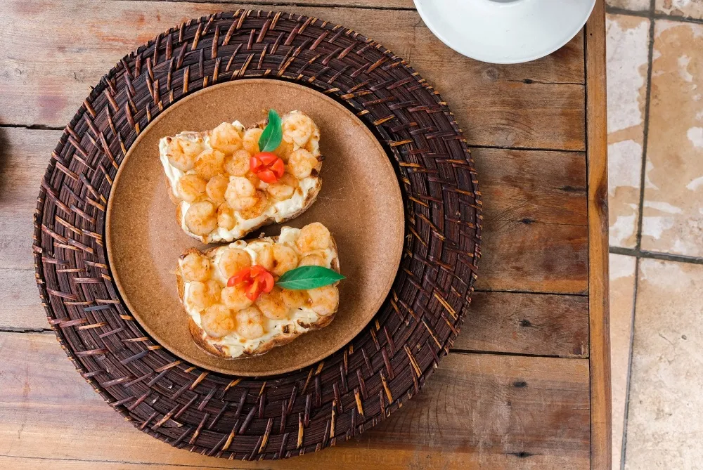 O Duo de Camarão Crocante é feito com duas fatias de pão artesano, camarão puxado na manteiga e cream cheese maçaricado