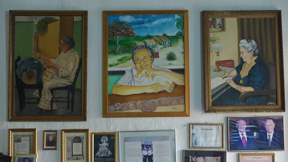 Parede de fotografias e pinturas na casa de Mestra Assunção, com um autorretrato ao centro: arte e memória juntas