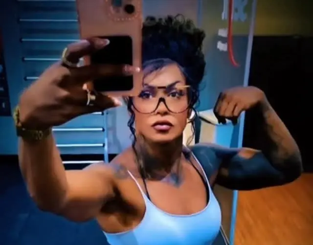 Mulher forte com óculos, cabelos escuros presos para trás, vestindo regata branca, mostrando força ao tirar selfie em ambiente interno.