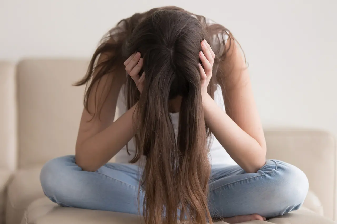 Imagem de jovem de calça jeans e blusa branca com mãos na cabeça, que está abaixada, e cabelo encobrindo o rosto