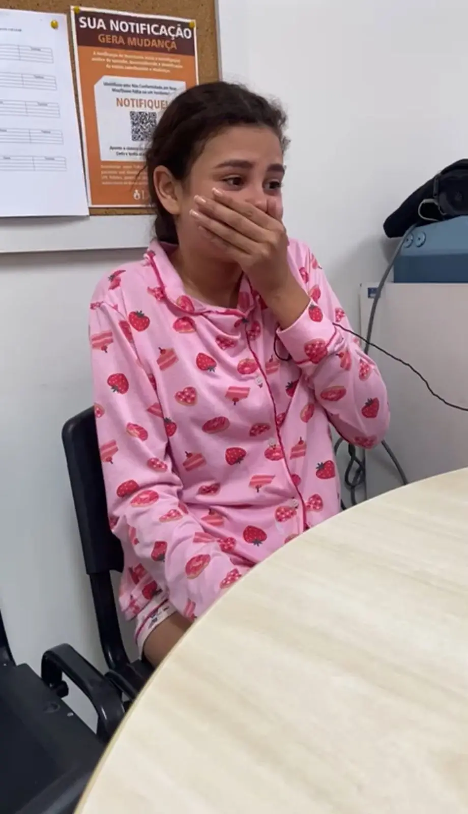 Na imagem, uma jovem, possivelmente Laís Vitória, sentada em uma cadeira preta em uma sala de escritório, vestindo um pijama rosa estampado com morangos e pedaços de bolo. Ela cobre a boca com a mão em uma expressão de surpresa e emoção, com lágrimas nos olhos, sugerindo que acabou de receber uma notícia impactante. Um mural ou quadro de avisos com papéis e um cartaz que diz 